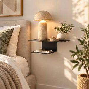 May include: A black metal wall-mounted floating shelf with two tiers. The top tier holds a beige mushroom-shaped lamp and a small round bowl. The bottom tier holds a rectangular smartphone. The shelf is mounted on a light-coloured wall.