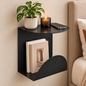 May include: A matte black metal wall-mounted shelf featuring a flat top surface and a curved lower storage section. The shelf holds a green potted plant in a white ribbed ceramic pot, a lit candle in a dark glass jar, a smartphone, and two books.