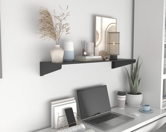 May include: A black floating shelf with a white vase, a blue vase, a book, a lamp, and other decorative items. The shelf is mounted on a white wall above a white desk with a laptop, a magazine, a phone, and a cup.