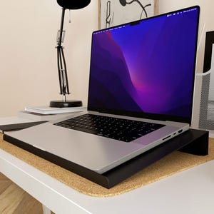 Può includere: Un laptop argentato con tastiera nera è posizionato su un supporto nero. Lo schermo mostra una sfumatura viola e blu. Il supporto è su una superficie di sughero e la scrivania è bianca.