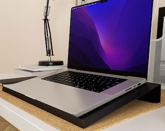 May include: A silver laptop with a black keyboard sits on a black laptop stand. The screen displays a purple and blue gradient. The stand is on a cork surface, and the desk is white.