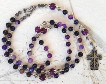 May include: A rosary necklace with a cross pendant and beads in shades of purple, black, and silver. The beads are interspersed with silver-toned metal accents. The cross pendant has an ornate design.