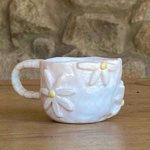 Peut inclure: Une tasse en céramique faite à la main avec un glaçage rose clair et des décorations de marguerites. La poignée est rayée de jaune et de blanc. La tasse est posée sur une surface en bois.