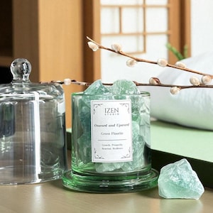 May include: A glass cloche and a cylindrical glass jar filled with green fluorite crystals. The jar has a label that reads "IZEN STUDIO" and "Onward and Upward Green Fluorite". A single fluorite crystal sits beside the jar. A sprig of pussy willow is in the background.
