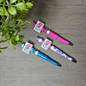 May include: Three decorative pens with square charms featuring floral designs and the text "With all things are possible." The pens have pink, blue, and purple barrels, accented with beads. A green plant is in the background.