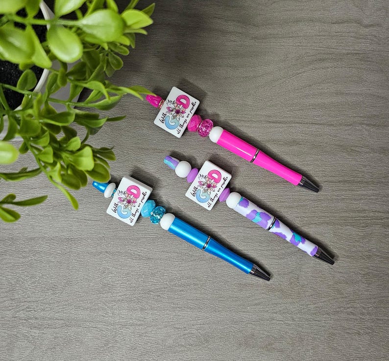 May include: Three decorative pens with square charms featuring floral designs and the text "With all things are possible." The pens have pink, blue, and purple barrels, accented with beads. A green plant is in the background.