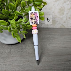 May include: White ballpoint pen with a decorative top featuring a rectangular charm with the text "Sometimes I just have to let God fix it, cuz if I fix it I am going to jail." The pen has pink, orange, and patterned beads.