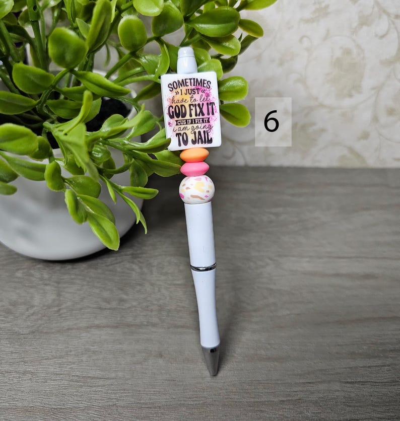 May include: White ballpoint pen with a decorative top featuring a rectangular charm with the text "Sometimes I just have to let God fix it, cuz if I fix it I am going to jail." The pen has pink, orange, and patterned beads.