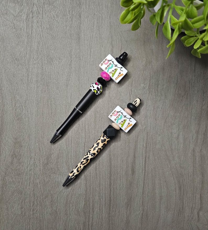 May include: Two decorative pens with black barrels and beaded accents. One pen has a leopard print pattern, while the other has a black and white spotted design. Both pens feature a white rectangular charm with the word "just" and colorful designs.