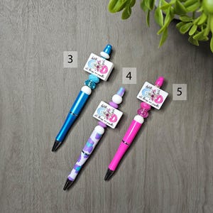 May include: Three decorative pens with unique designs. One is blue, one has a purple and white pattern, and the third is pink. Each pen has a square charm with the text "With God all things are possible" and floral accents.