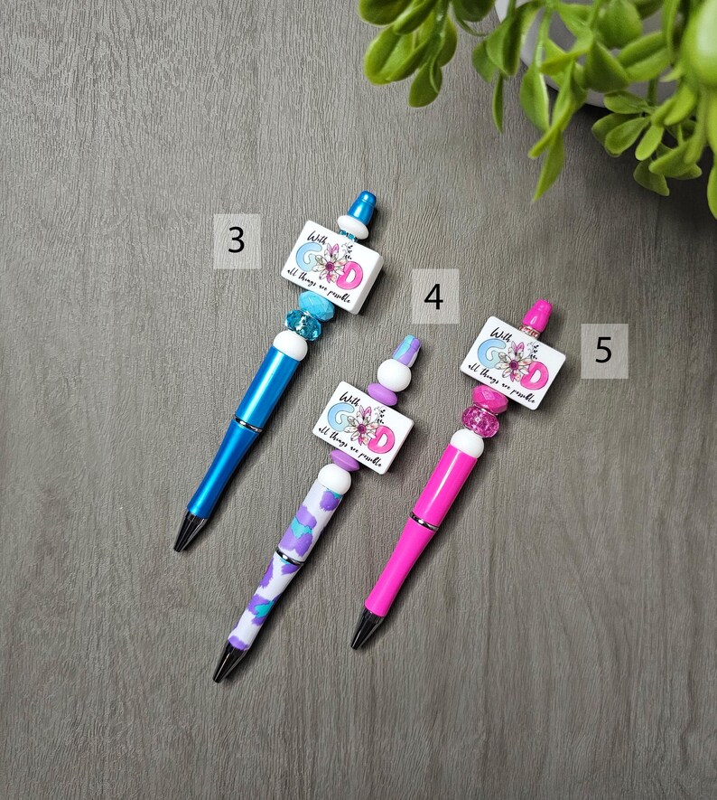 May include: Three decorative pens with unique designs. One is blue, one has a purple and white pattern, and the third is pink. Each pen has a square charm with the text "With God all things are possible" and floral accents.