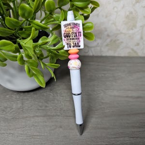 May include: White ballpoint pen with a decorative top featuring a rectangular charm with the text "Sometimes I just have to let God fix it..." and colorful beads. The pen has a silver tip and a curved design.