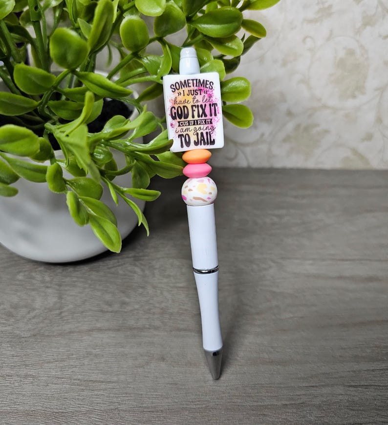 May include: White ballpoint pen with a decorative top featuring a rectangular charm with the text "Sometimes I just have to let God fix it..." and colorful beads. The pen has a silver tip and a curved design.