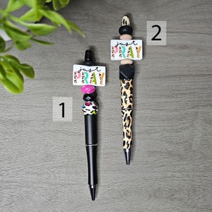 May include: Two decorative pens with black barrels. One pen has a pink, black, and white beaded design, and the other has a leopard print design. Both pens have a white rectangular charm with the words "just pray".