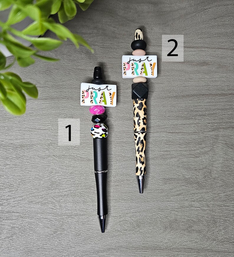 May include: Two decorative pens with black barrels. One pen has a pink, black, and white beaded design, and the other has a leopard print design. Both pens have a white rectangular charm with the words "just pray".
