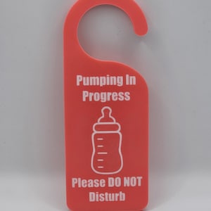 May include: A coral-colored door hanger with a curved top and a rectangular bottom. White text reads "Pumping In Progress" above a baby bottle graphic and "Please DO NOT Disturb" below. The hanger is designed to be placed on a door handle.