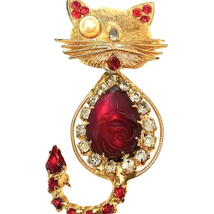 Op de afbeelding: Goudkleurige kattenbroche met rode en heldere kristallen accenten. De kattenkop heeft rode kristallen en een imitatie parel oog. De body heeft een grote, druppelvormige rode steen met een rozenontwerp, omringd door heldere kristallen.