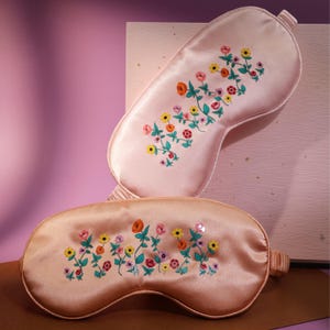 Könnte beinhalten: Zwei rosa Satin-Schlafmasken mit Blumenstickerei. Die Masken haben einen hellrosa Hintergrund mit bunten Blumen und grünen Blättern.
