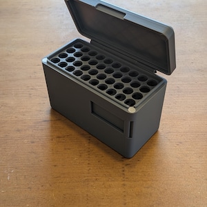 以下が含まれることがあります： 開いた蓋が付いた、濃い灰色の長方形の収納ボックス。内部には複数の円形のコンパートメントがあります。ボックスはマットな素材でできており、木製の表面に置かれています。蓋はヒンジで取り付けられており、ハンドルが付いています。