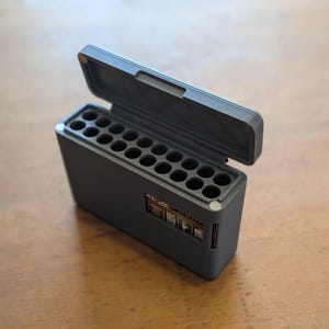 May include: A dark grey rectangular case with an open lid, showing multiple circular compartments. The case has a small digital display on the side. The case is on a wooden surface.