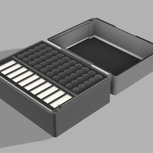 May include: A grey storage box, open to reveal internal compartments. One section features rectangular slots, another has circular indentations, and a third is empty. The lid is open, showing a black interior.