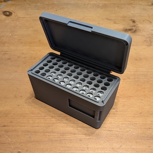 Pode incluir: Uma caixa de armazenamento cinza retangular com uma tampa aberta. O interior apresenta uma grade de orifícios circulares. A caixa foi projetada para guardar pequenos objetos cilíndricos. A caixa é feita de material fosco.