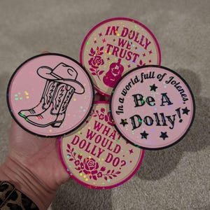 May include: Four circular stickers with a holographic glitter finish. One features black cowboy boots and a hat. The others have pink text: IN DOLLY WE TRUST with a guitar and rose, Be A Dolly! with stars, and WHAT WOULD DOLLY DO? with a rose design.