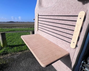 May include: A wooden shelf attached to the inside of a vehicle door. The shelf is made of light brown wood and has a rounded edge. The shelf is supported by two wooden brackets. The shelf is attached to the door with screws. The door is covered in a light gray fabric. The shelf is in a parked vehicle with a blue sky in the background.