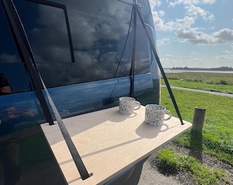 May include: A wooden table attached to the back of a blue van with two black and white mugs on top. The table is supported by two black straps.