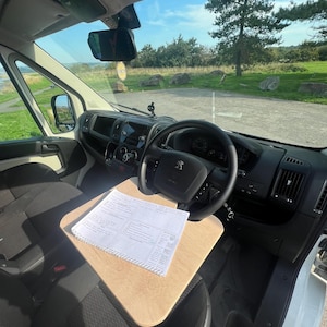 May include: A white van interior with a wooden desk mounted on the dashboard. A notebook with a checklist is on the desk. The van is parked in a grassy area with a tree in the background.