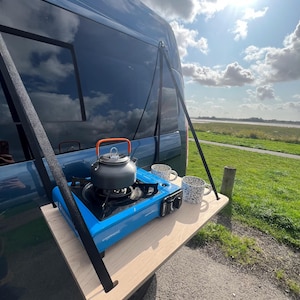 May include: A portable camping kitchen set up on a wooden platform attached to the back of a blue van. The set includes a blue camping stove, a silver kettle, and two mugs.