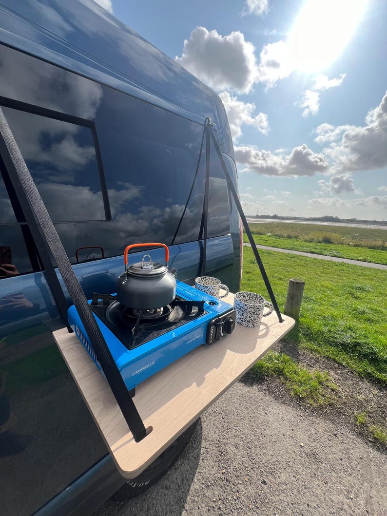 May include: A portable camping kitchen set up on a wooden platform attached to the back of a blue van. The set includes a blue camping stove, a silver kettle, and two mugs.
