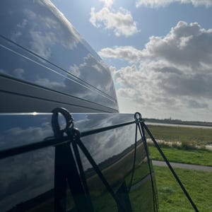 May include: A black van with a black privacy screen attached to the back window. The screen is held in place by two black straps with metal clips. The van is parked in a grassy area with a blue sky and white clouds in the background.