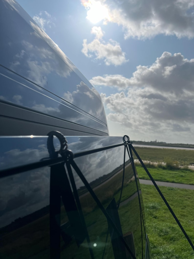 May include: A black van with a black privacy screen attached to the back window. The screen is held in place by two black straps with metal clips. The van is parked in a grassy area with a blue sky and white clouds in the background.