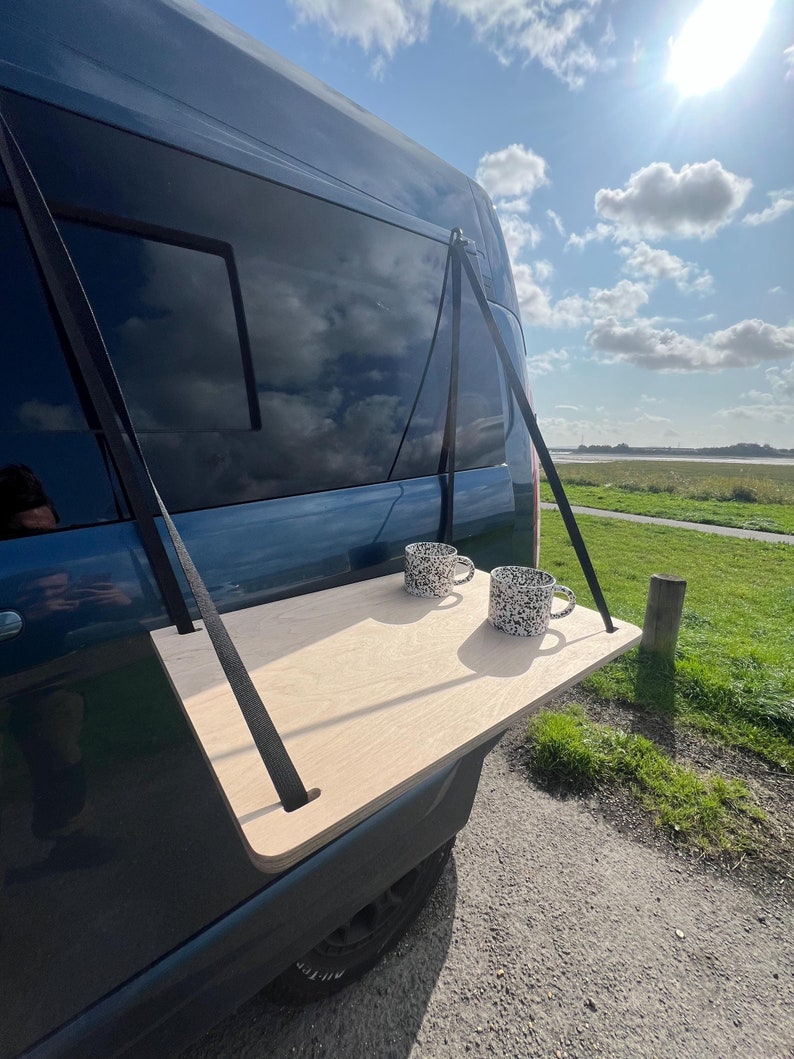 May include: A wooden table attached to the back of a blue van with two black and white mugs on top. The table is supported by two black straps.