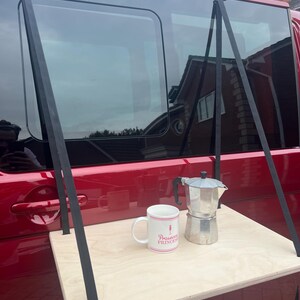 May include: A wooden table with black straps attached to the top. The table is hanging from the back of a red van. A white mug with the text "Prosecco Princess" and a silver coffee pot are on the table.