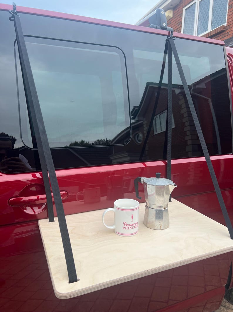 May include: A wooden table with black straps attached to the top. The table is hanging from the back of a red van. A white mug with the text "Prosecco Princess" and a silver coffee pot are on the table.