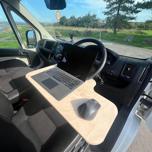 May include: A white van with a laptop and mouse on a wooden desk attached to the passenger seat. The van is parked in a grassy area with a tree in the background.
