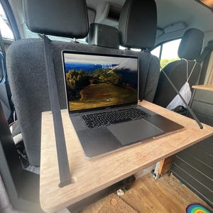 May include: A laptop computer with a screen displaying a landscape scene is placed on a wooden table that is attached to the back of two car seats. The table is held in place by two black straps.