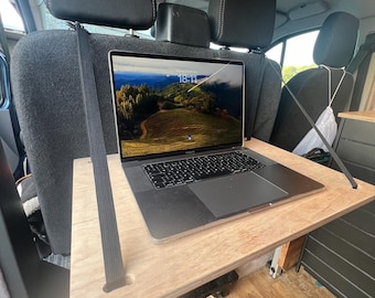 May include: A laptop computer with a screen displaying a landscape scene is placed on a wooden table that is attached to the back of two car seats. The table is held in place by two black straps.