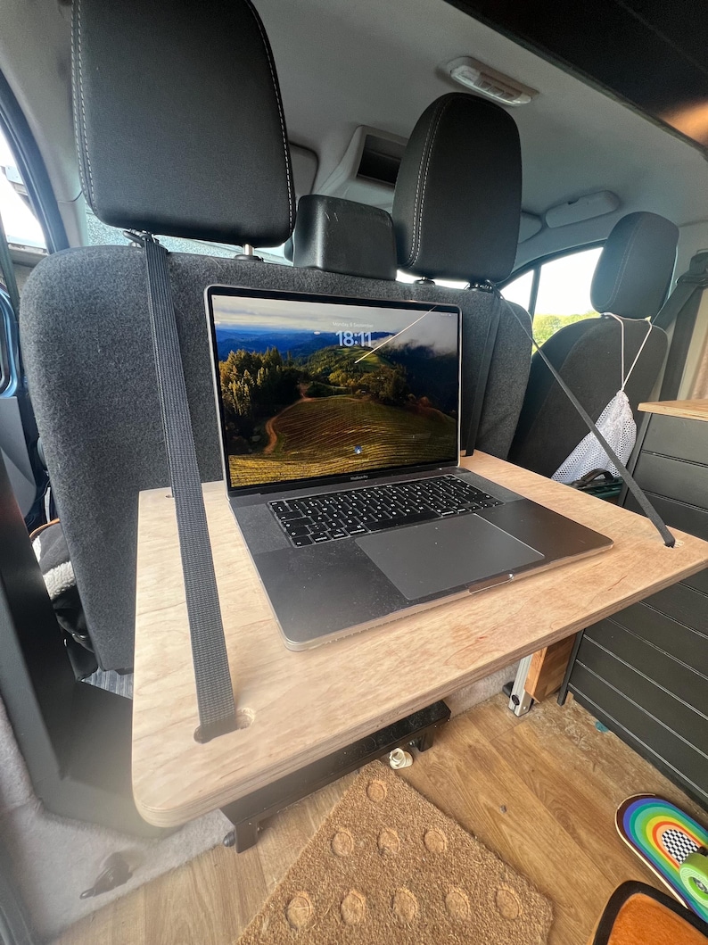 May include: A laptop computer with a screen displaying a landscape scene is placed on a wooden table that is attached to the back of two car seats. The table is held in place by two black straps.