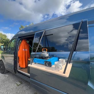 May include: A blue van with a wooden table attached to the back door. The table has a small blue gas stove, a kettle, and two mugs on it.