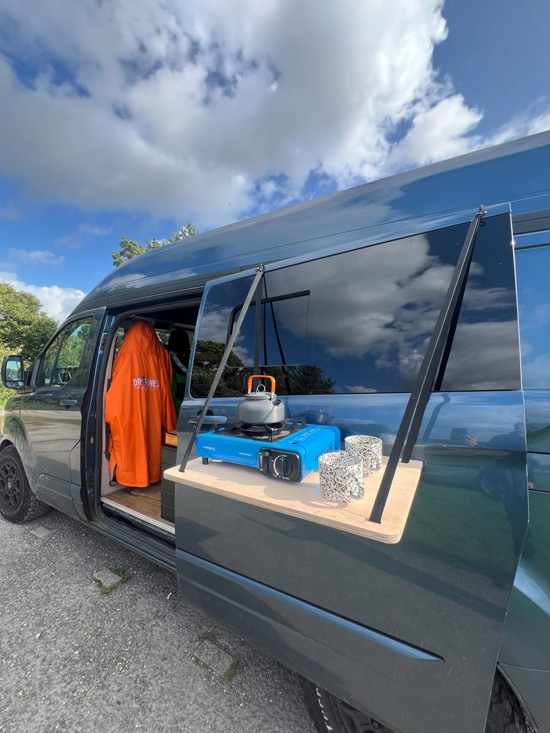 May include: A blue van with a wooden table attached to the back door. The table has a small blue gas stove, a kettle, and two mugs on it.