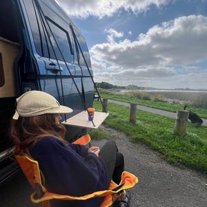 May include: A person sits in an orange and yellow folding chair with a blue fleece jacket and a beige hat. The chair is in front of a blue van with a table attached to the side. The table has a small orange and blue cup on it. The van is parked in a grassy area with a cloudy sky in the background.