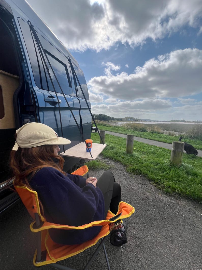 May include: A person sits in an orange and yellow folding chair with a blue fleece jacket and a beige hat. The chair is in front of a blue van with a table attached to the side. The table has a small orange and blue cup on it. The van is parked in a grassy area with a cloudy sky in the background.