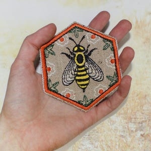 May include: Hexagon-shaped embroidered patch featuring a detailed bee design. The bee is yellow and black, set against a beige background with an orange border. The patch has a decorative floral pattern.