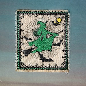 May include: A rectangular fabric patch with a frayed edge featuring an embroidered green witch flying on a broomstick. The design includes black bats, a yellow moon, and concentric circular stitching on a beige background with a black border.