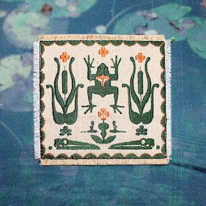 May include: A square fabric patch with a cream-coloured base featuring dark green embroidery of a central frog flanked by two cattail plants. The design includes small orange floral accents and a scalloped dark green border with frayed edges.