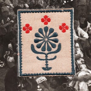 May include: A square patch with a beige background and a dark blue border. The patch features a central dark blue floral design with red flower accents. The background is a woven fabric, and the patch is likely an embroidered emblem.