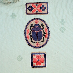 May include: Three embroidered patches with blue, red, and tan designs. The top patch is rectangular with a floral pattern. The middle patch is oval with a scarab beetle. The bottom patch is square with a four-petal flower.